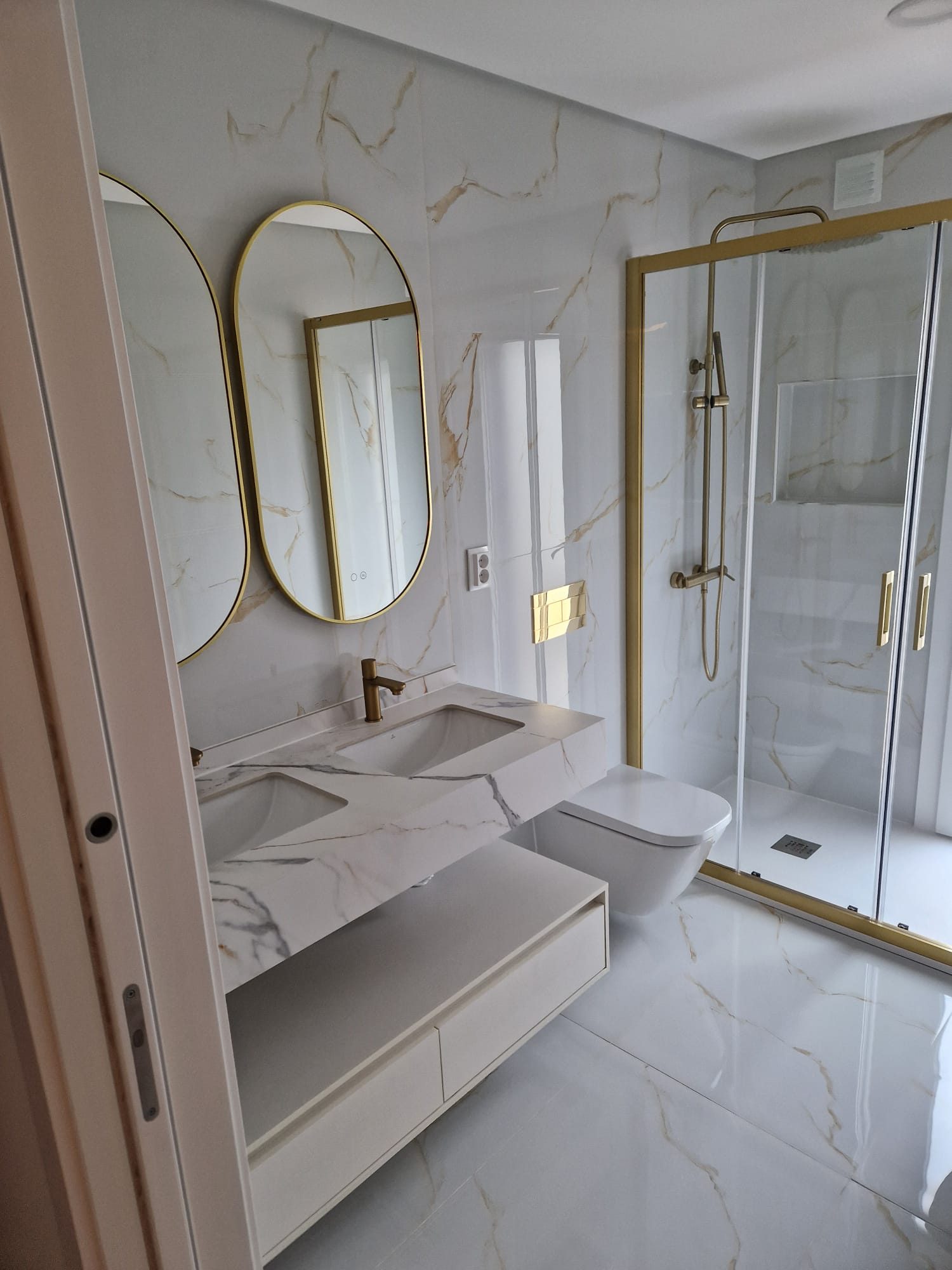 Casa de banho revestida a mármore claro, com espelho oval de aro dourado e base de duche ao nível do pavimento